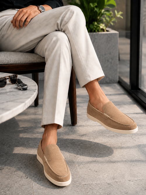 A person seated on a chair wearing light beige trousers and tan slip-on loafers with white soles in a modern indoor setting.