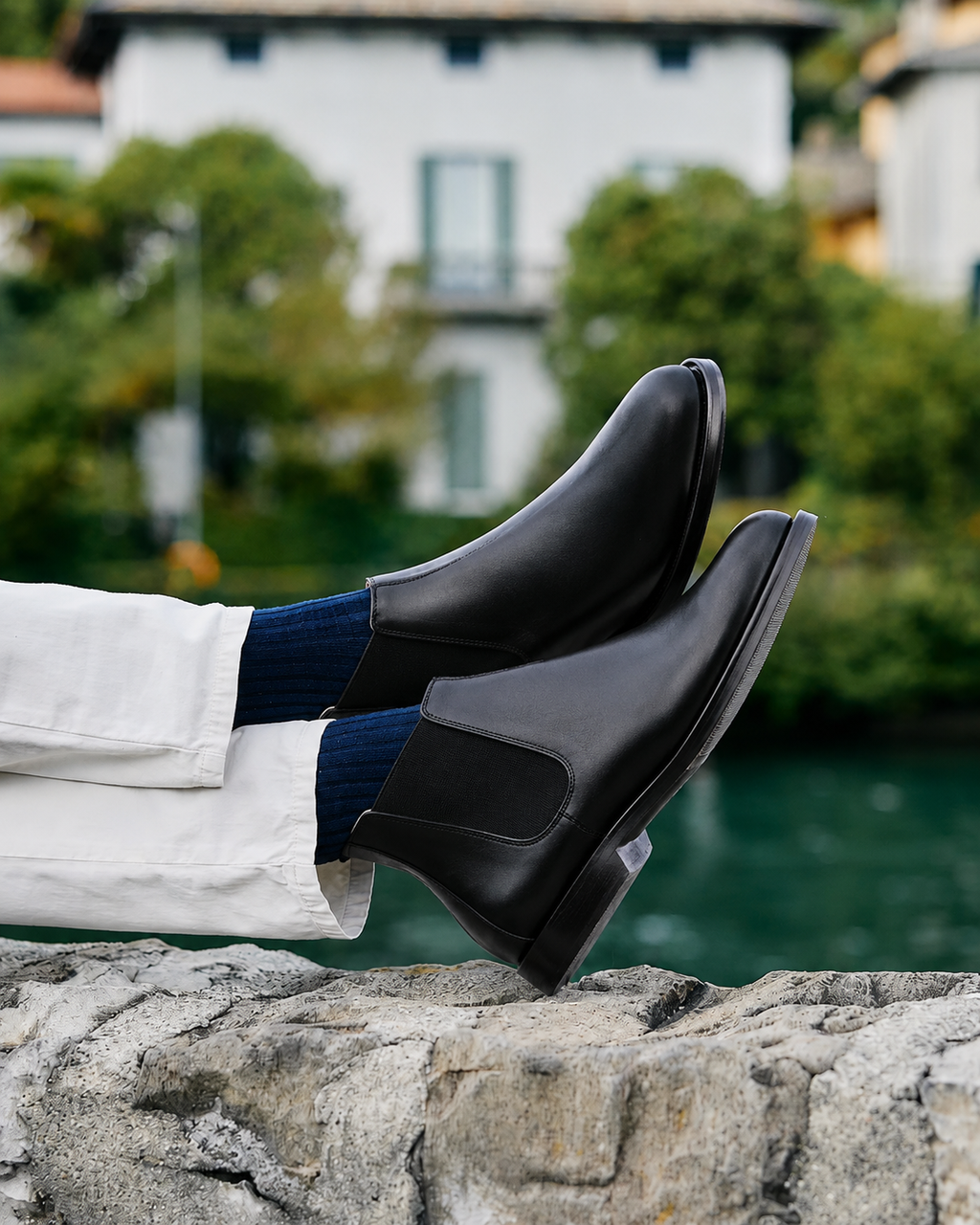 A pair of black Chelsea boots worn with white trousers and navy socks, shown in a lifestyle setting with a stone wall and a blurred background of water and a white building.