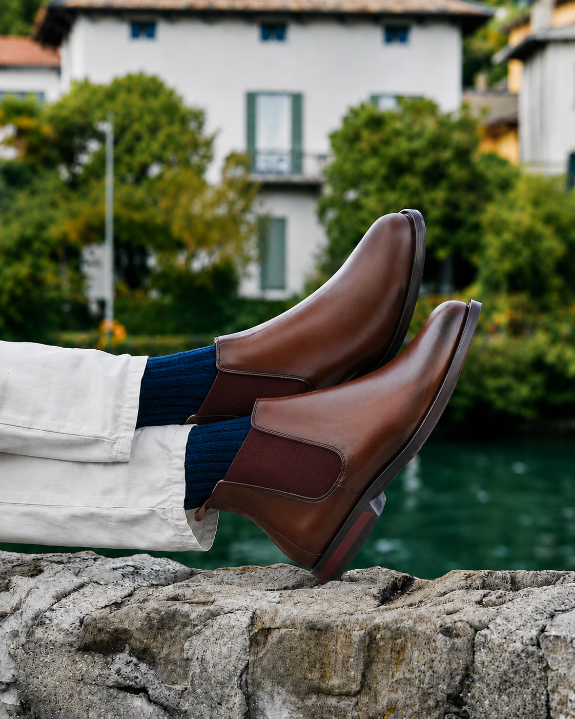 A pair of brown leather Chelsea boots worn with white trousers and navy blue socks, resting on a stone ledge with a scenic lake and white building in the background.