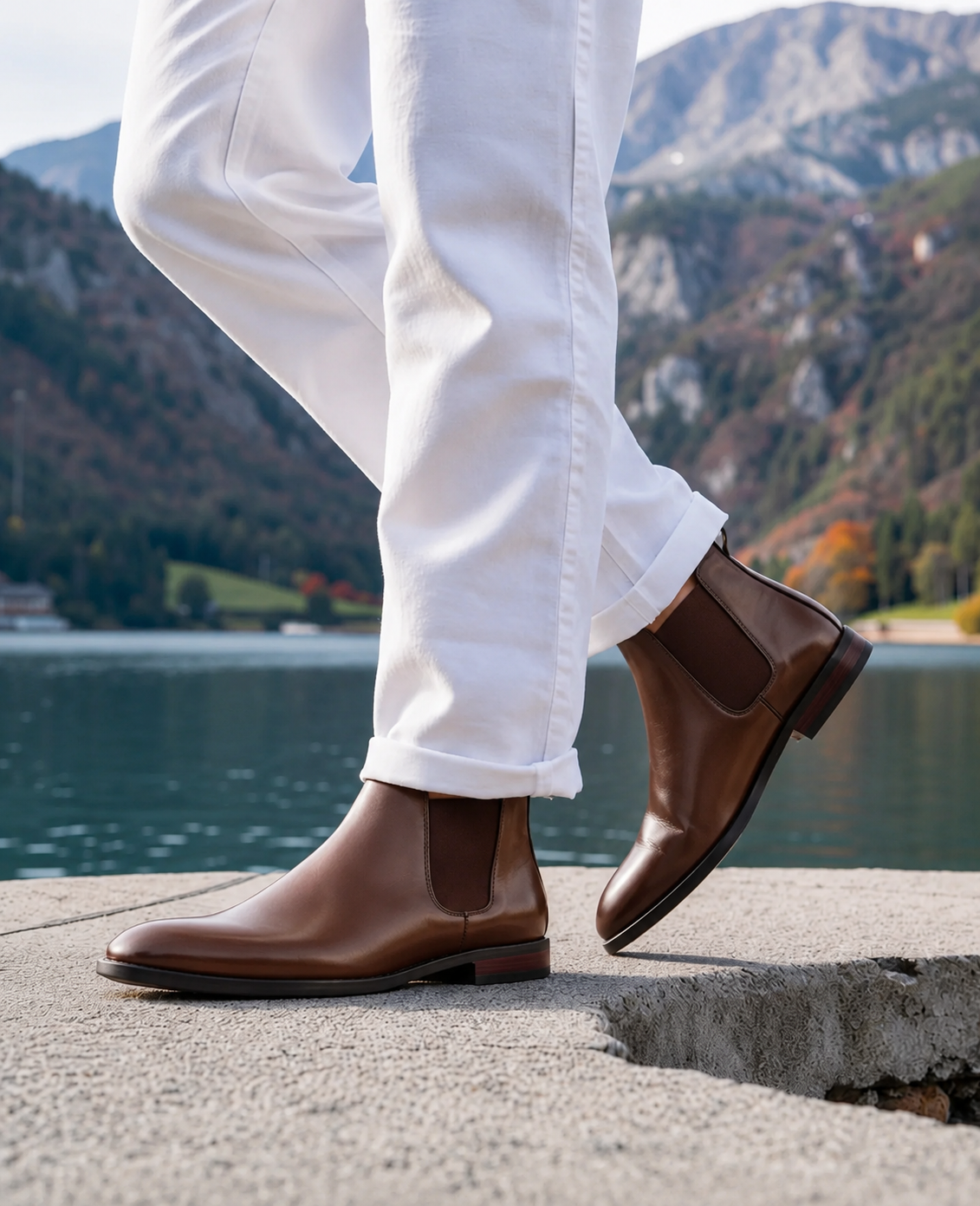A person wearing brown leather Chelsea boots with white pants, standing on a concrete ledge with a scenic mountain lake in the background.