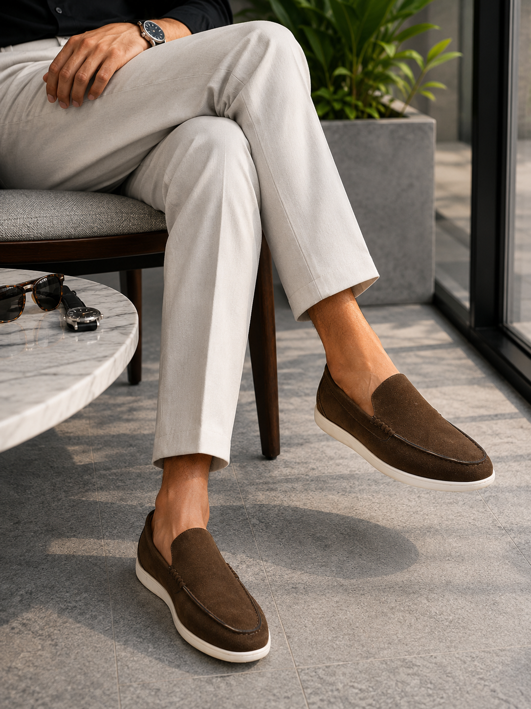 A person seated on a chair wearing brown suede slip-on loafers with white soles, paired with light beige trousers in a modern indoor setting.