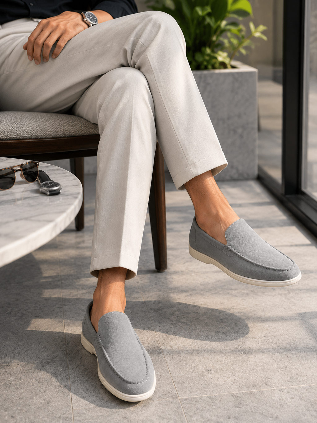 A person seated on a chair wearing light gray slip-on loafers with off-white soles, paired with light beige trousers in a modern indoor setting.