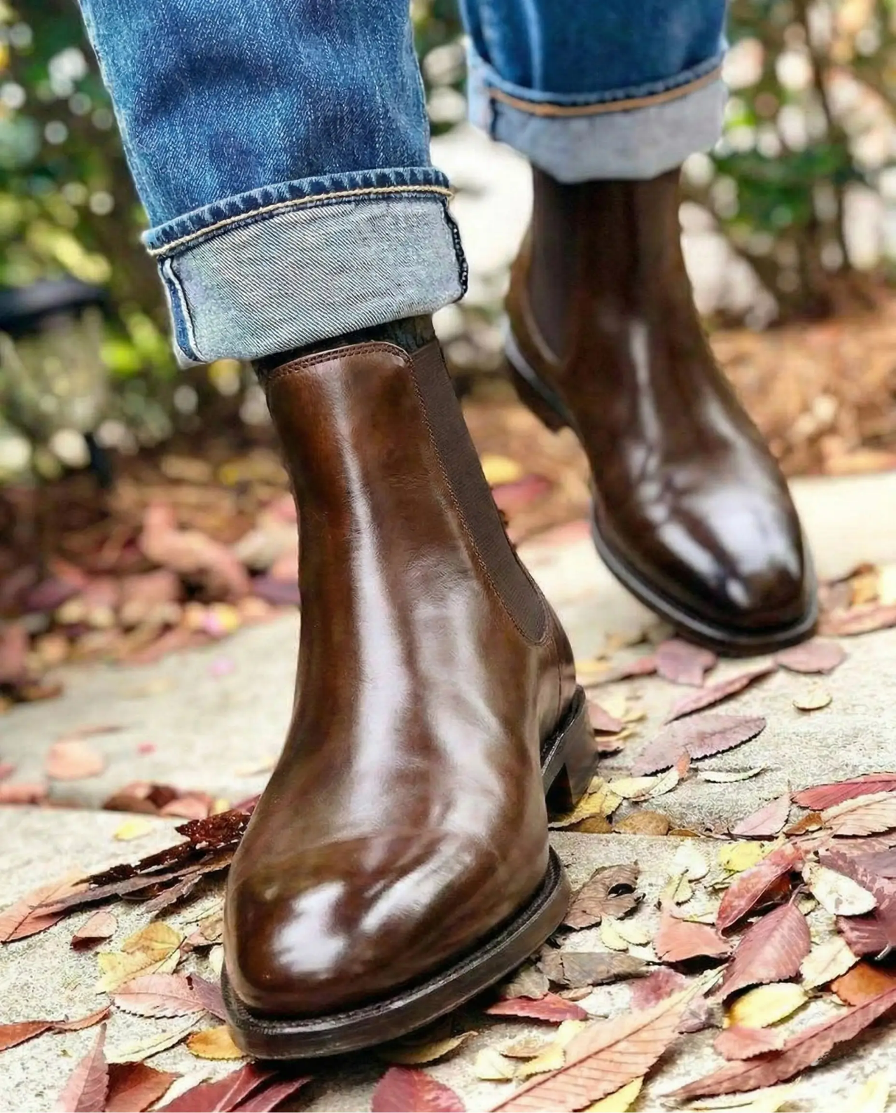 Brown leather boots worn with rolled-up jeans on a leaf-covered ground.