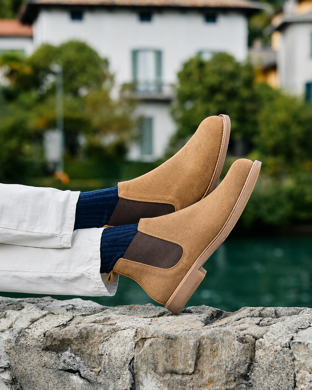 A person wearing tan suede Chelsea boots with dark brown elastic side panels, paired with white trousers and navy socks, sitting on a stone ledge with a blurred outdoor background of water and a white building.