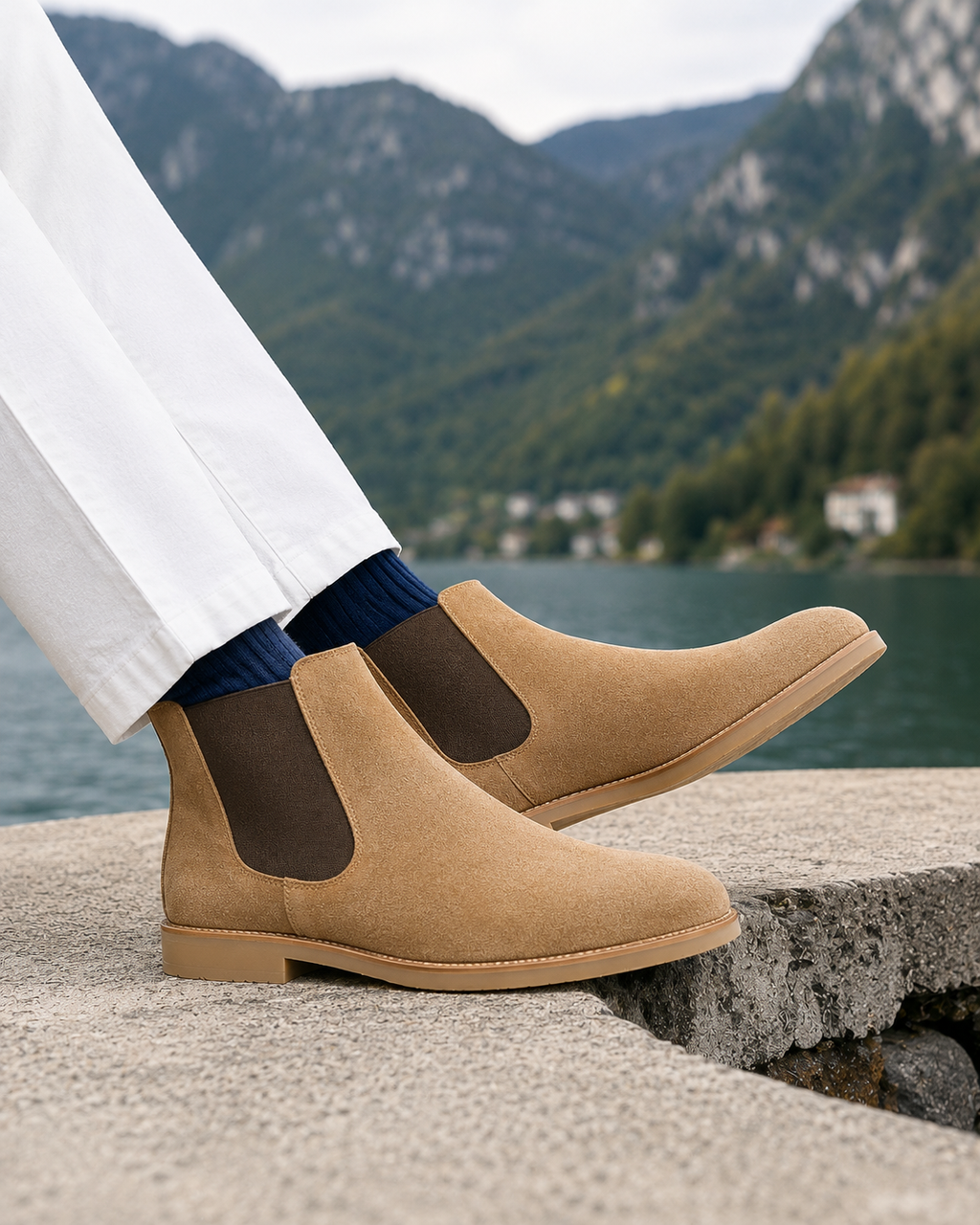 A person wearing tan suede Chelsea-style ankle boots with dark brown elastic side panels, paired with white trousers and navy socks, sitting on a stone ledge with a mountain lake landscape in the background.