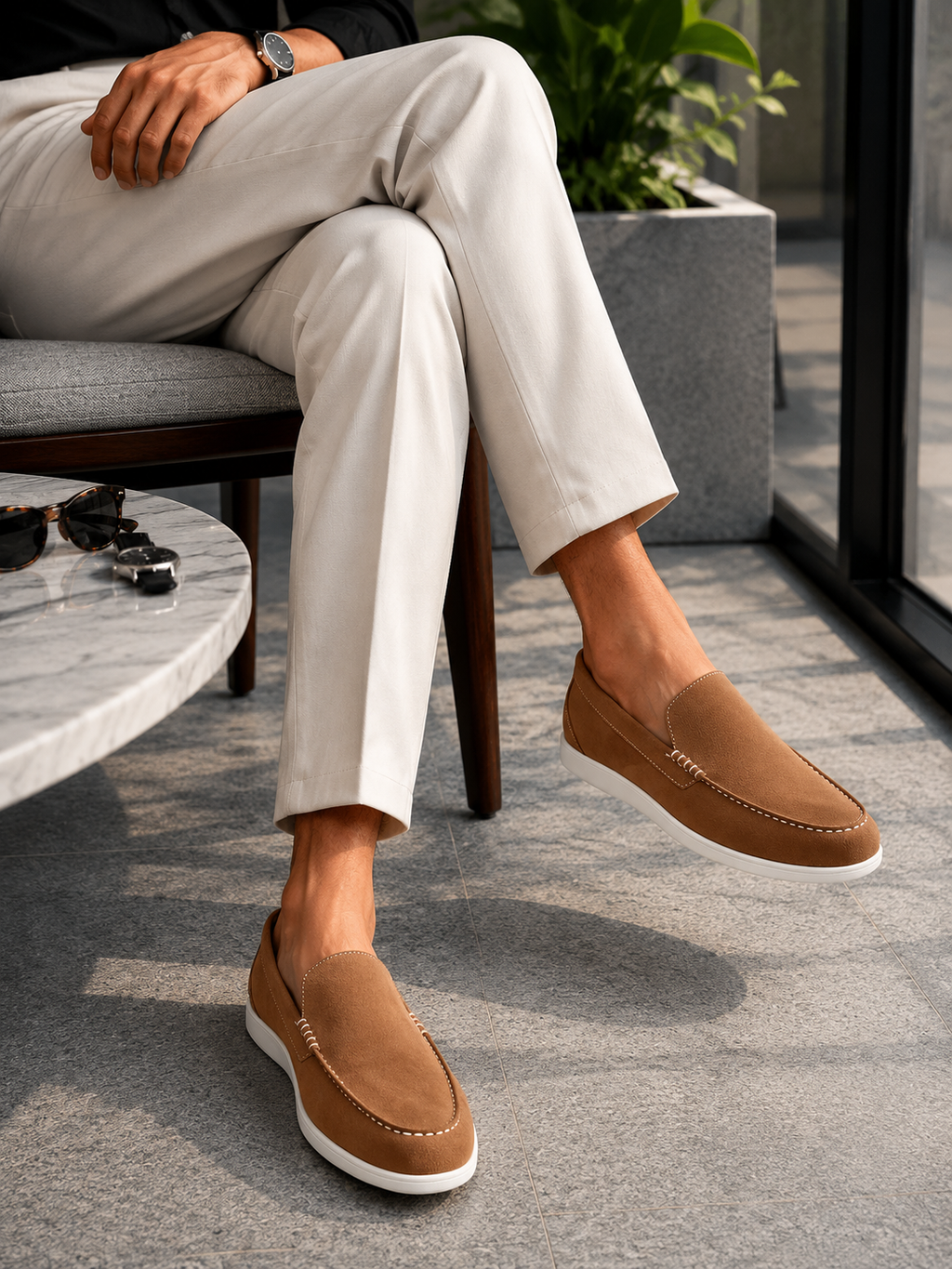 A person seated on a chair wearing tan slip-on loafers with white soles, paired with light beige trousers in a modern indoor setting.